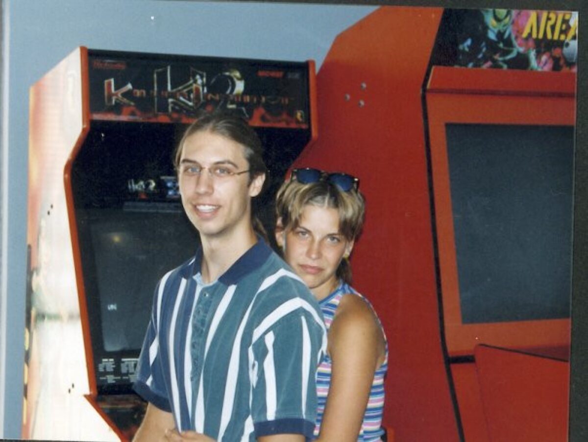 Interior of the original Area 51 arcade, 1990s. Two large orange arcade cabinets against a wall, with two young people standing beside them.